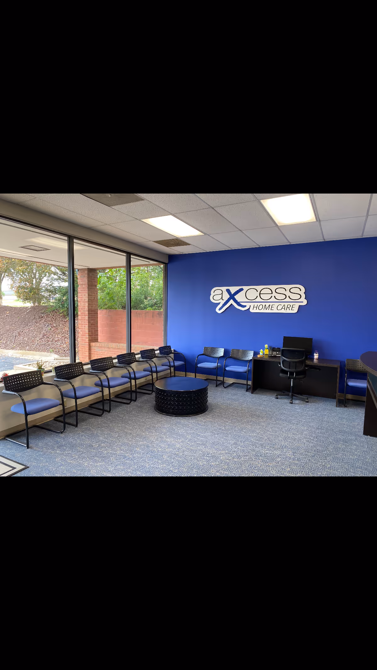 Interior waiting area with blue wall logo, chairs along windows, and a central table.