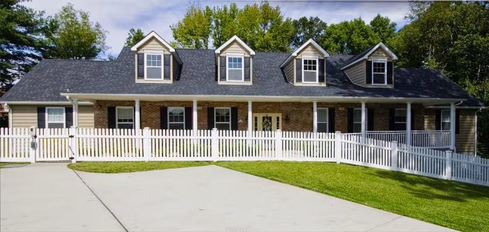 Front exterior of a house-like senior living facility with a white picket fence and green lawn.