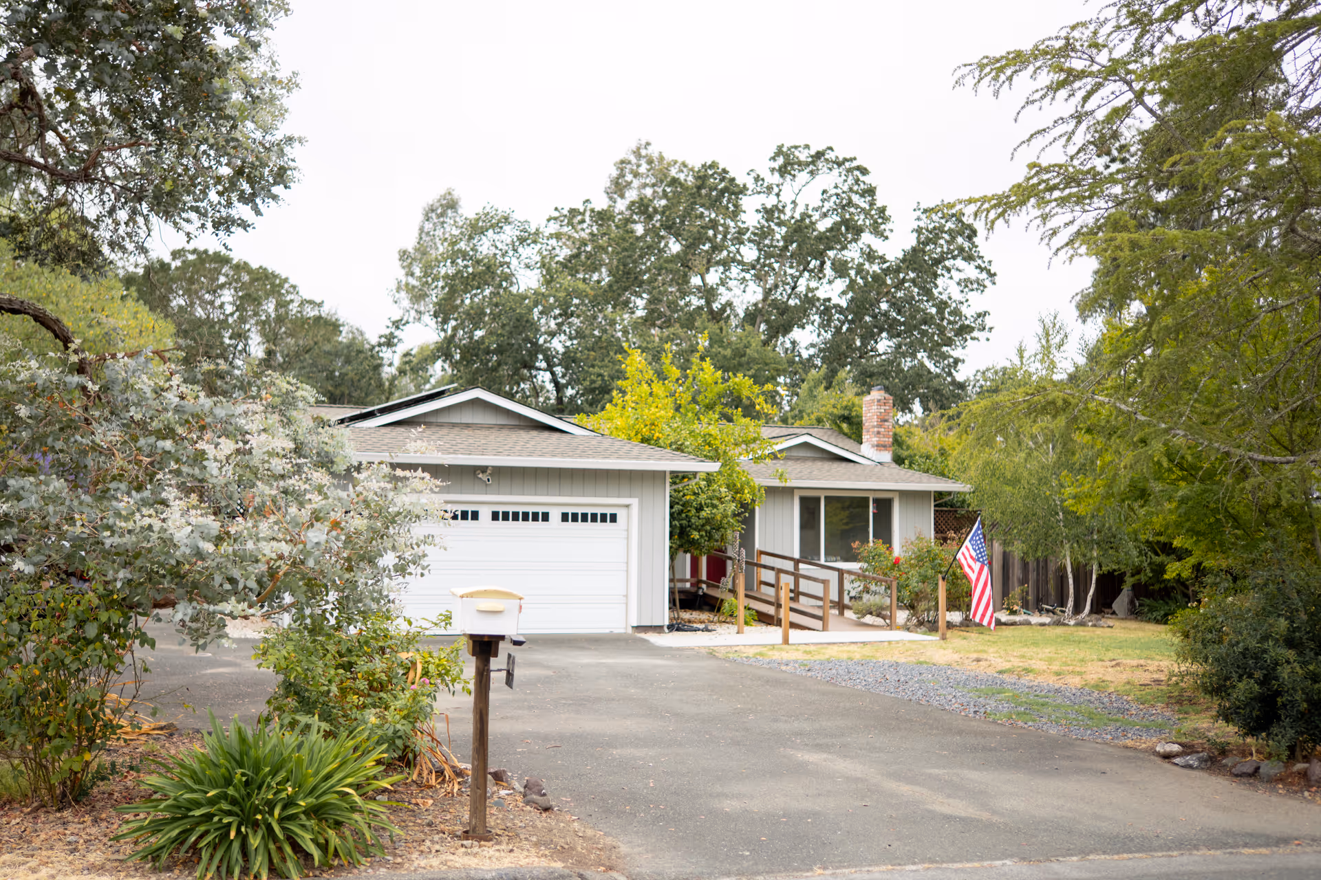 Single-story house with ramp, driveway, and American flag in a lush front yard.