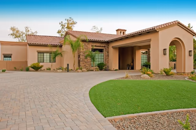 Exterior view of a single-story building with a tiled roof and beige stucco walls, featuring a covered entrance with an archway, surrounded by a paved driveway, green lawn, and desert landscaping with small palm trees and shrubs under a clear blue sky.