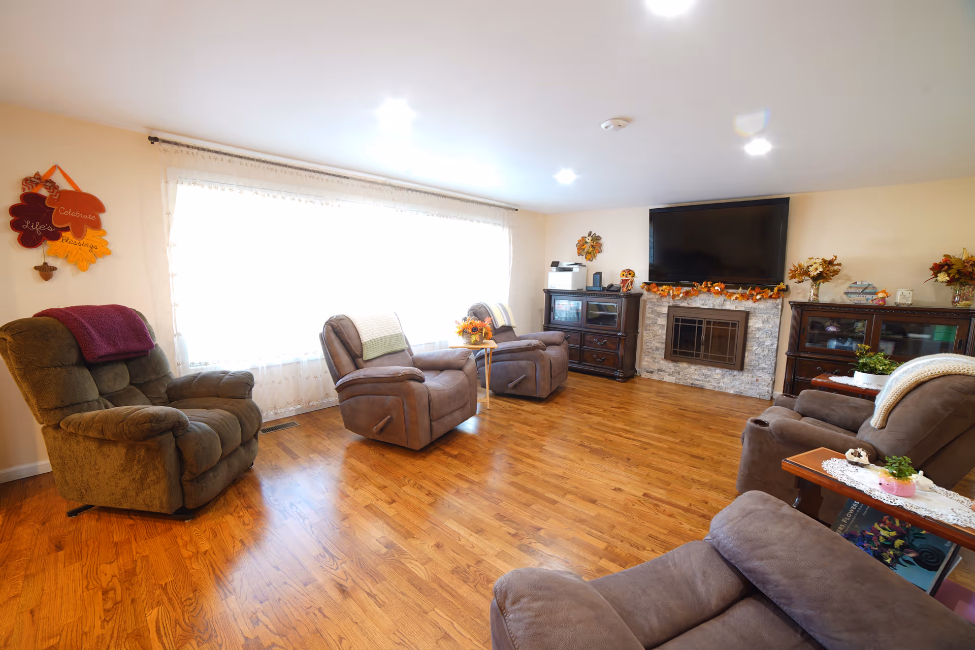 Bright senior living lounge with multiple recliners, a large TV, and a decorative fireplace.