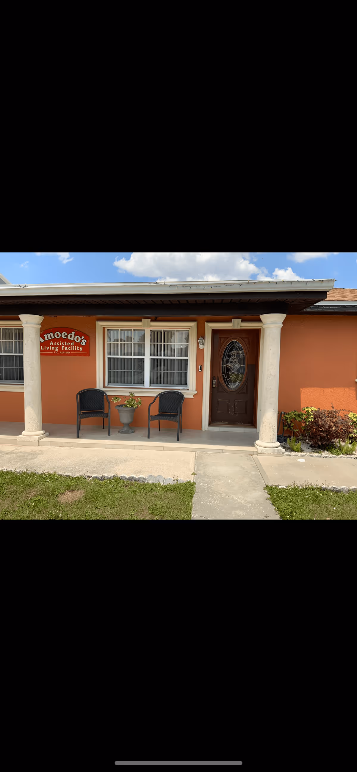 Front exterior view of Amoedo's Assisted Living Facility showing an orange building with two white columns supporting a small porch roof. There are two black chairs and a planter with flowers on the porch, a window with white curtains, and a brown door with an oval decorative glass panel. A red sign with white text reads 'Amoedo's Assisted Living Facility Lic. AL11160'.