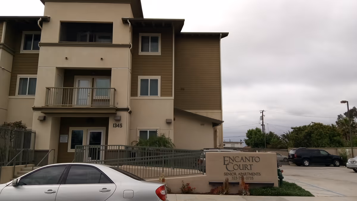 Three-story tan apartment building with Encanto Court Senior Apartments sign near a parking lot.
