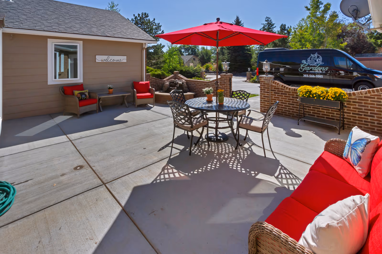 Outdoor patio at a senior living facility with red-cushioned seating, a round metal table and umbrella, and a branded van visible nearby.