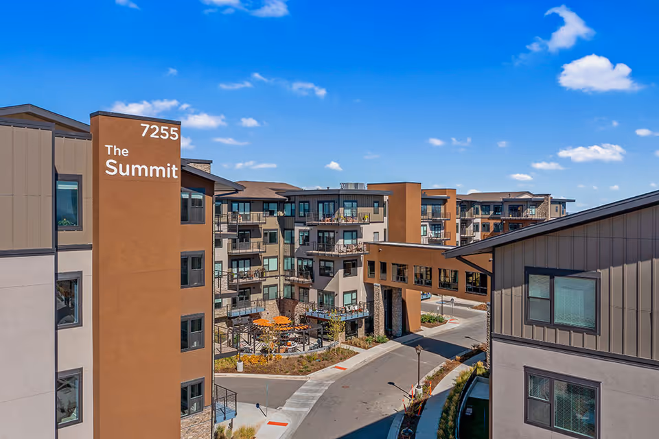 Exterior view of a modern multi-story senior living facility named The Summit, featuring balconies, a connecting skybridge, landscaped areas, and a clear blue sky.