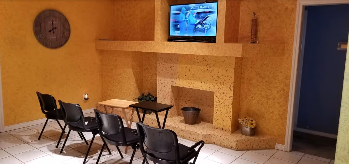 Indoor lounge with a mounted TV above a faux fireplace and a row of black chairs.