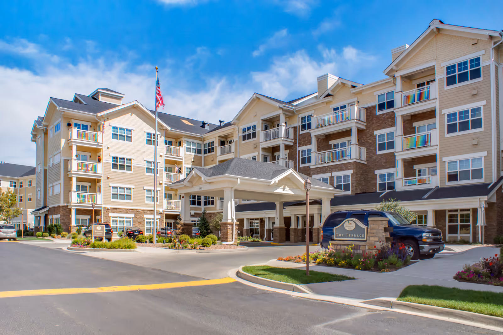Exterior view of a multi-story senior living facility building with beige and brown siding, multiple balconies, an American flag on a flagpole, a covered entrance, and a sign reading 'The Terrace' surrounded by landscaping and parked vehicles.