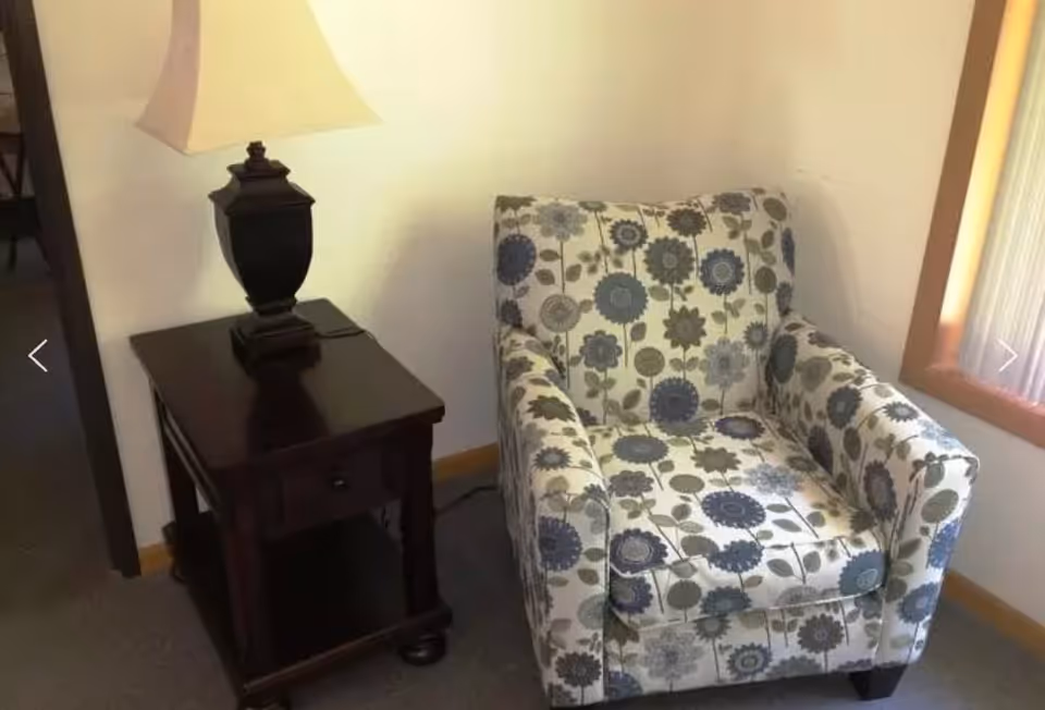 Patterned upholstered armchair next to a dark wooden side table with a lamp in a corner of a room.
