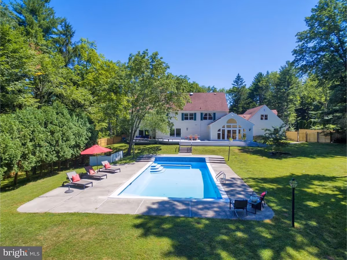 Outdoor view of a large white two-story house with a red roof, surrounded by green trees and a well-maintained lawn. In the foreground, there is a rectangular swimming pool with clear blue water, bordered by a concrete deck. Several lounge chairs with red cushions and a red umbrella are arranged on the left side of the pool, and a small seating area with two chairs and a table is on the right side. The sky is clear and blue.