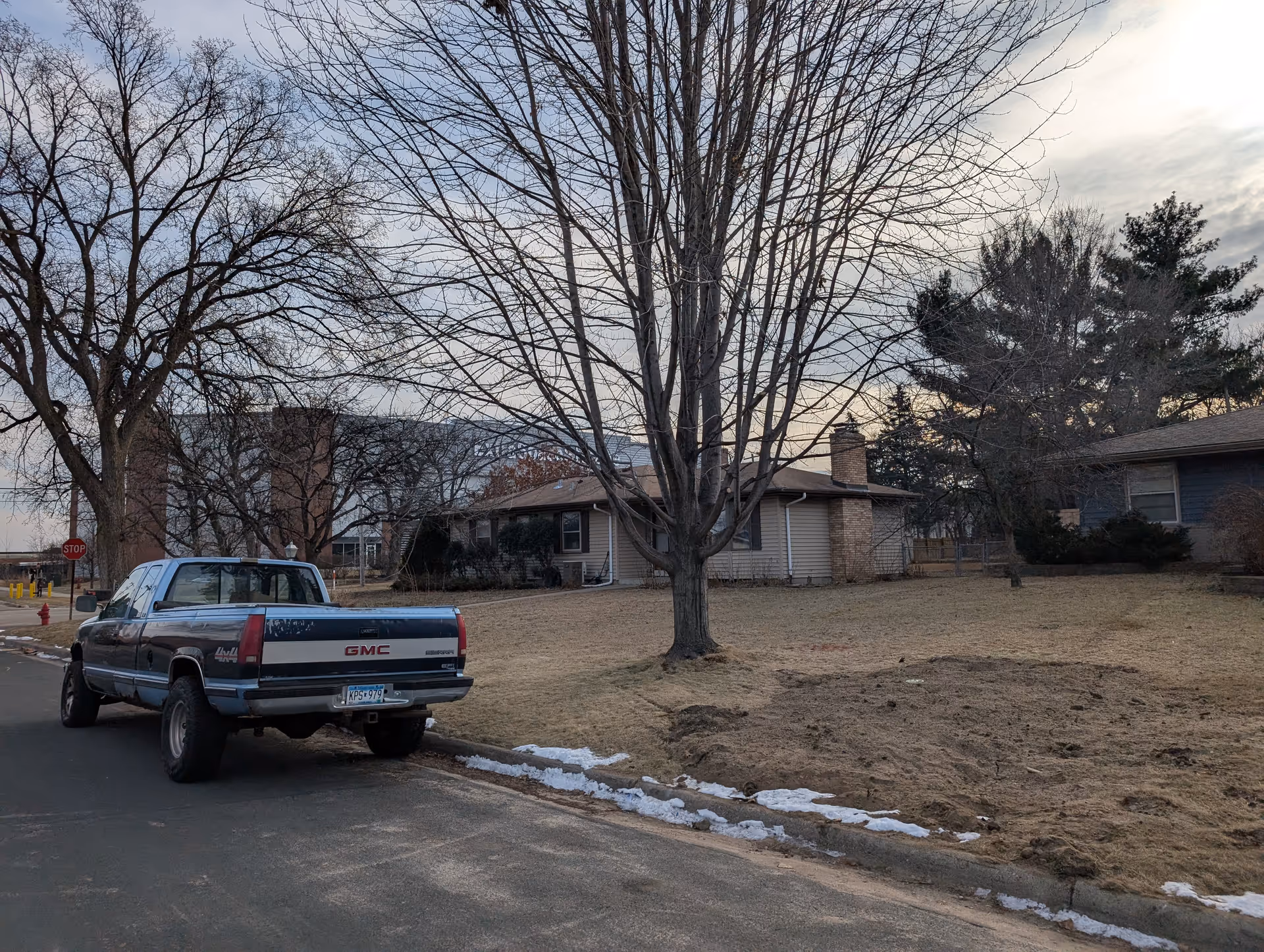 Suburban street with a parked GMC pickup truck, leafless trees, and single-story houses under a cloudy sky.