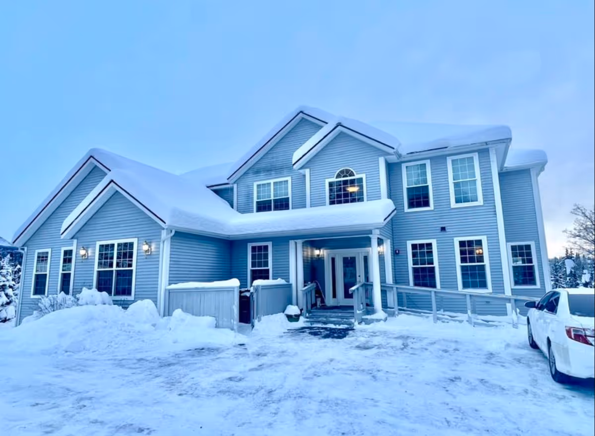 Snow-covered blue multi-story senior living facility exterior with a driveway and parked car.