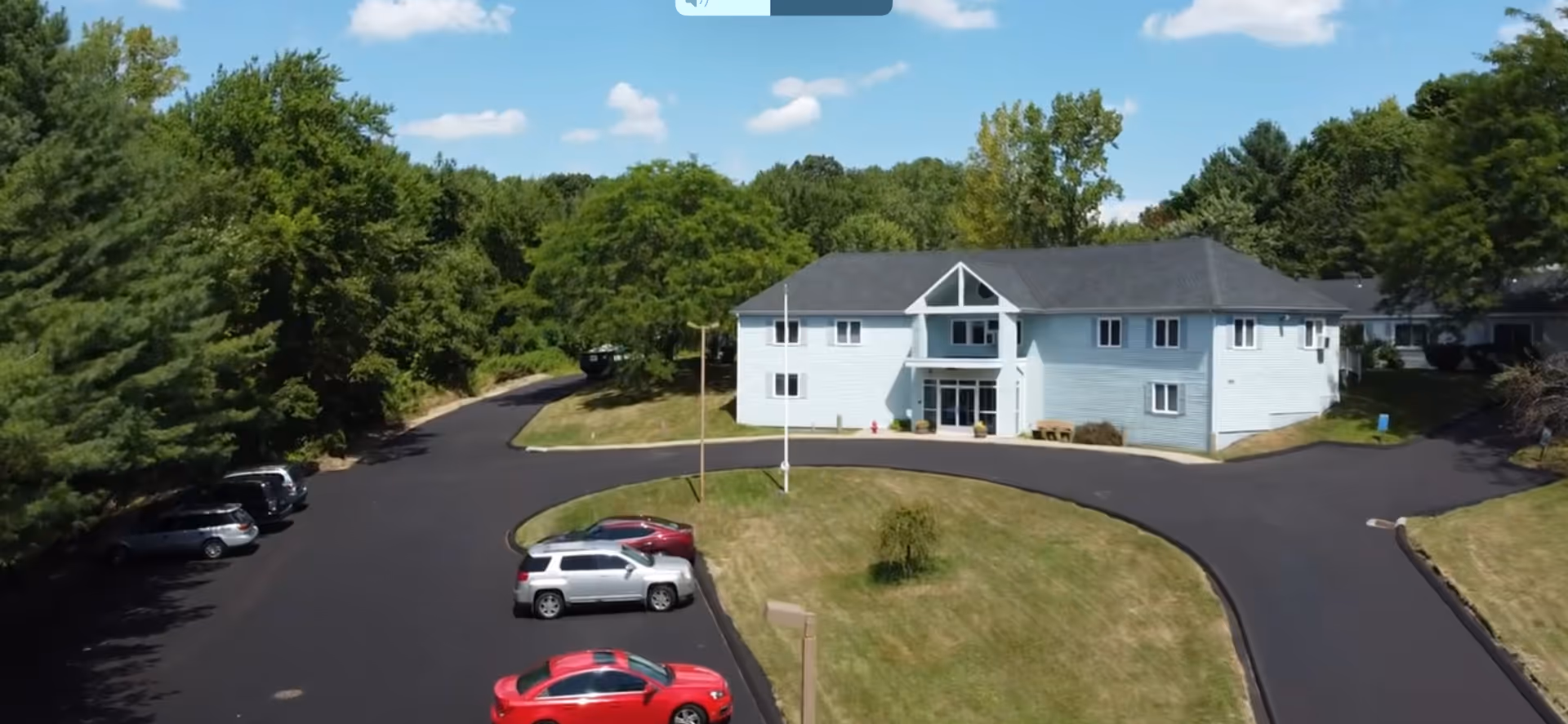 Aerial view of a light blue senior living building with a circular driveway and surrounding trees.