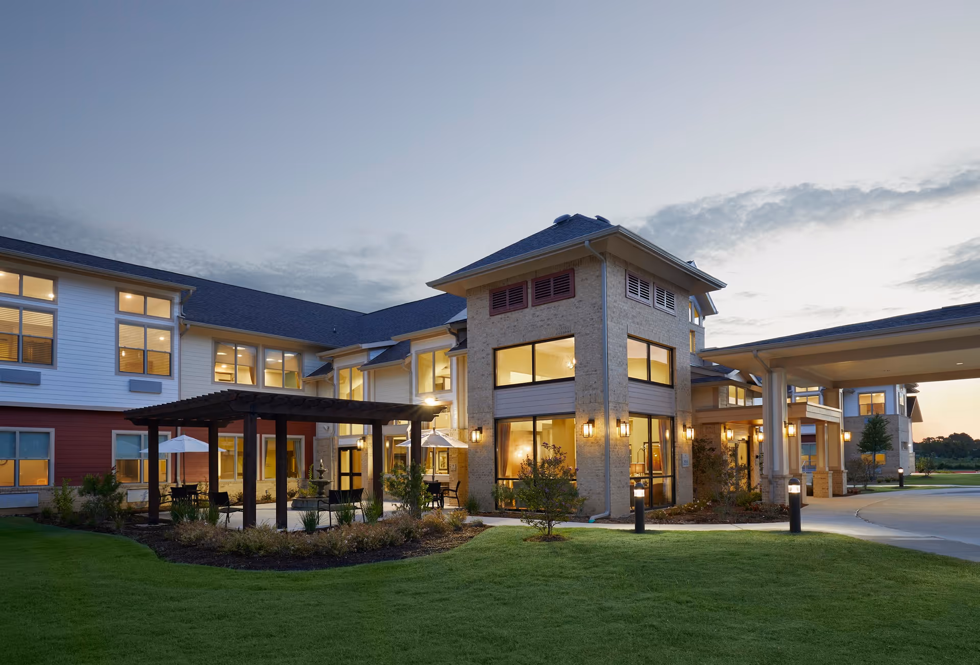 Exterior front view of a well-lit two-story senior living facility with a covered entrance, patio pergola, and landscaped lawn at dusk.