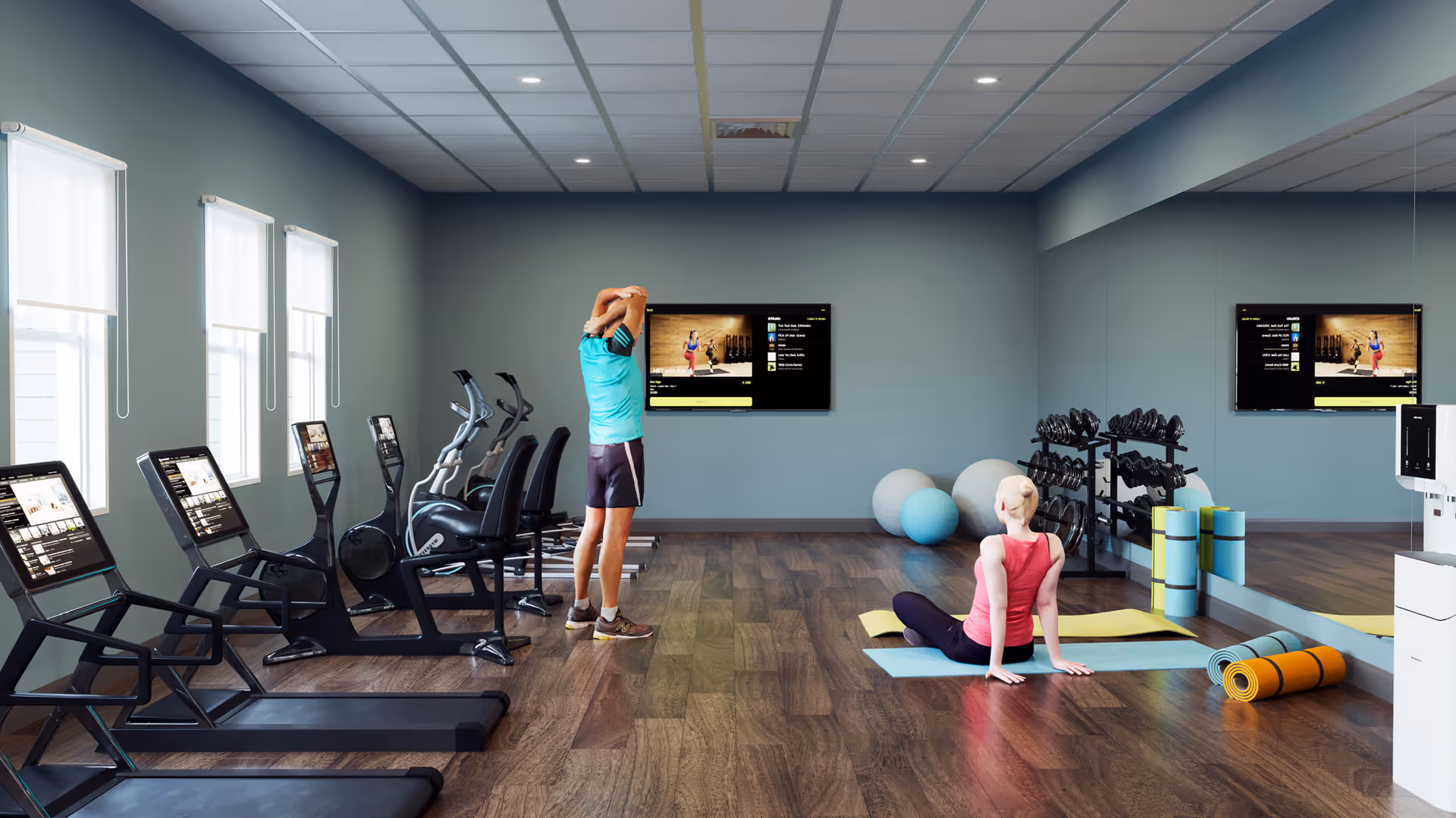 A fitness room with wooden flooring and light blue walls. There are several treadmills and exercise bikes lined up along the left side near windows. A man in athletic wear is stretching near the center of the room facing a wall-mounted TV. A woman in workout clothes is sitting on a yoga mat on the floor near exercise balls and foam rollers. A large mirror covers the right wall, reflecting the room and equipment.