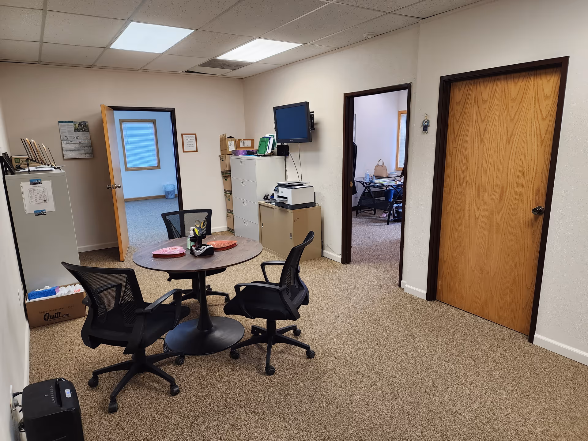 Office-style room with round table, chairs, filing cabinets, and a wall-mounted monitor.
