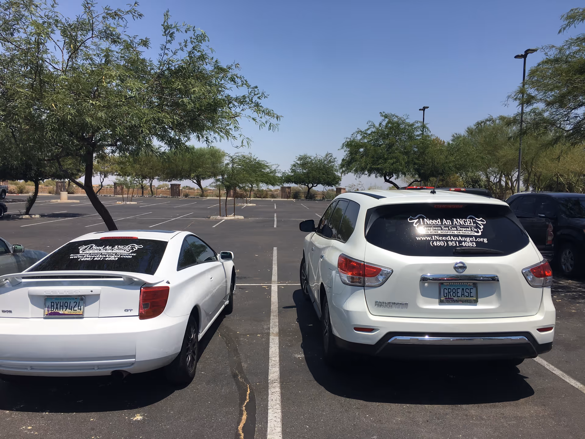 Two white cars parked in a sunny lot; rear window displays 'I Need An ANGEL' decal.