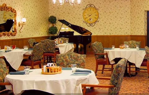 A dining room with round tables covered in white tablecloths, set with glasses, napkins, and condiments. Upholstered chairs with floral patterns surround the tables. A grand piano is positioned against the wall, which is decorated with floral wallpaper, a large clock, and a mirror. The room has warm lighting from wall sconces and a chandelier.