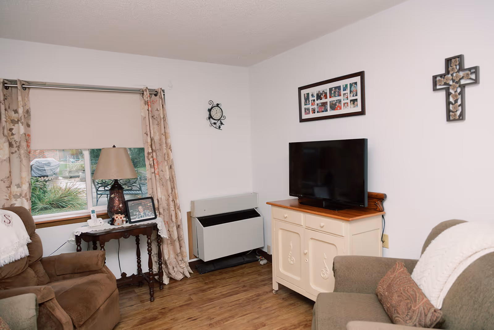 A cozy living room with a brown armchair and a green sofa, both with white throws. A small wooden table with a lamp, a phone, and a framed photo sits next to a window with floral curtains and a beige roller shade. A flat-screen TV is placed on a cream-colored cabinet with wooden top. The walls are white and decorated with a clock, a photo collage, and a decorative cross. The floor is wooden.