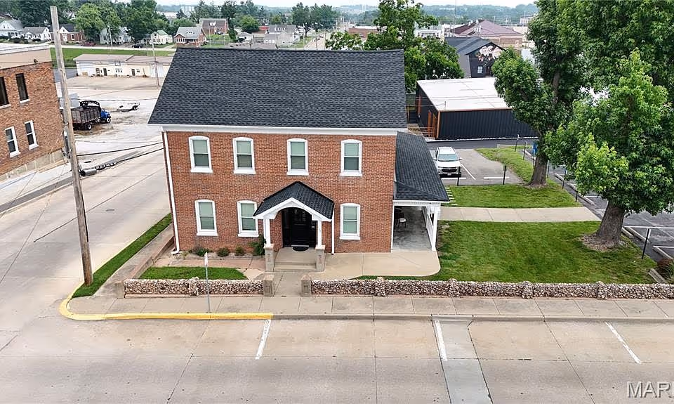 A brick, two-story building with a small front porch, stone wall, parking lot, and green lawn.