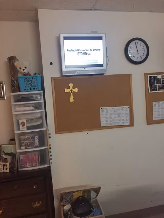 Interior wall with a mounted TV, clock, corkboard cross, and plastic storage tower.