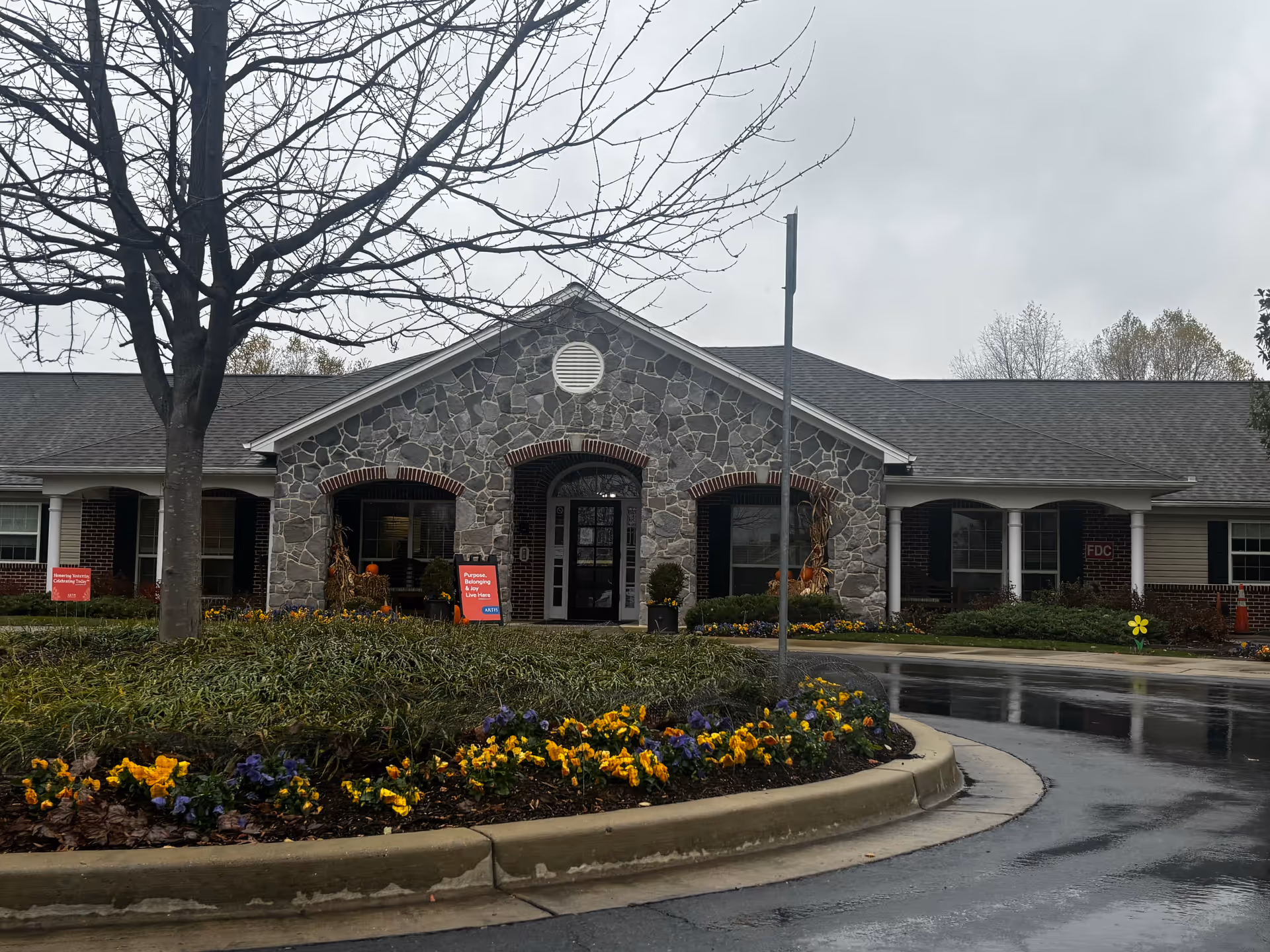 Front exterior of a single-story stone-faced memory care building with a circular driveway, landscaped flower beds, and a bare tree on a cloudy day.