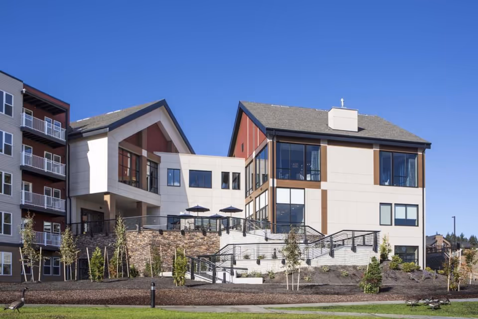 Front exterior of a modern multi-story senior living building with terraces, staircases, and landscaped grounds under a clear blue sky.