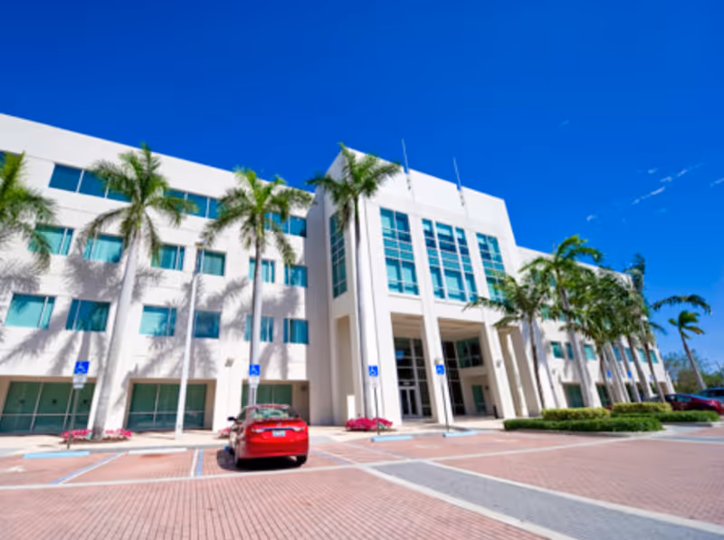 Exterior view of a modern white senior living facility with palm trees and parked cars.