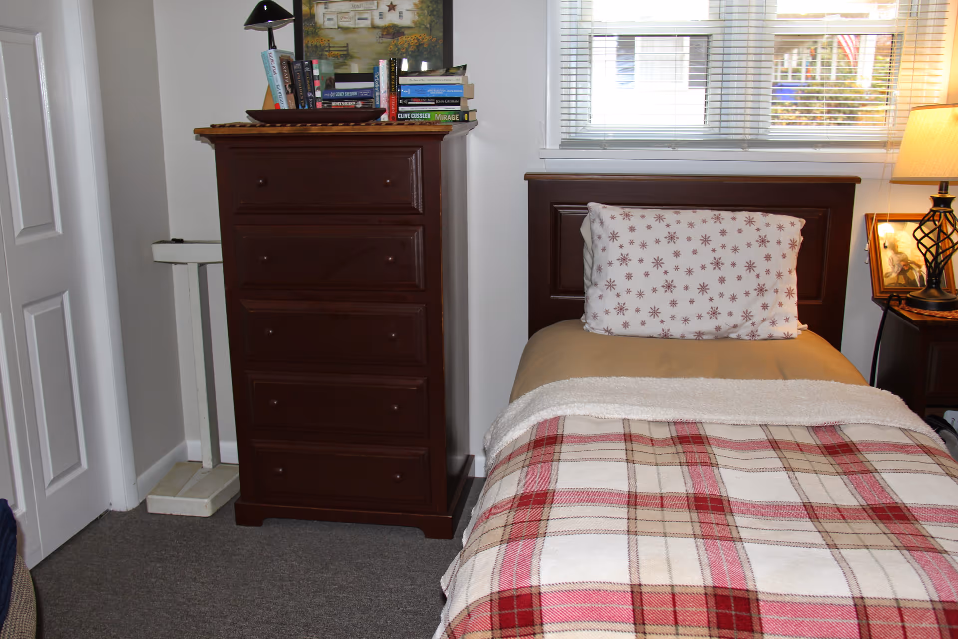 A bedroom with a single bed featuring a plaid blanket and a pillow with a star pattern. Next to the bed is a dark wooden nightstand with a lamp and a framed photo. To the left of the bed is a tall dark wooden dresser with several books and a lamp on top. The room has a window with blinds letting in natural light.
