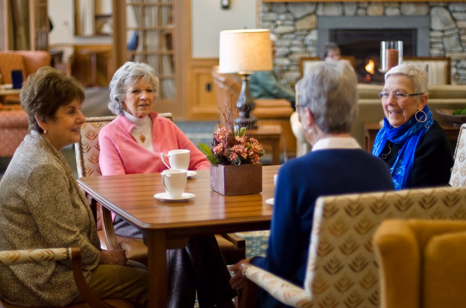 Four elderly women sitting around a wooden table in a cozy living room area with a stone fireplace in the background. They are engaged in conversation, with cups on the table and a floral centerpiece. The room has comfortable chairs and warm lighting.