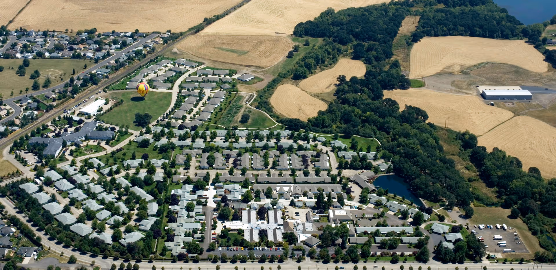 Aerial view of Mennonite Village senior living facility showing multiple buildings, roads, parking areas, green spaces, and surrounding farmland and wooded areas.