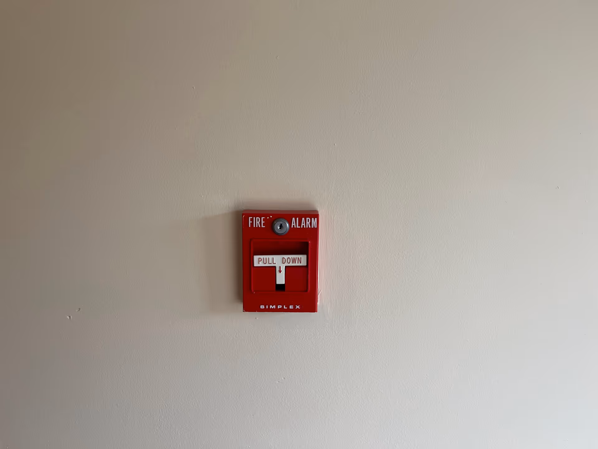 Red fire alarm pull station mounted on a plain beige wall.