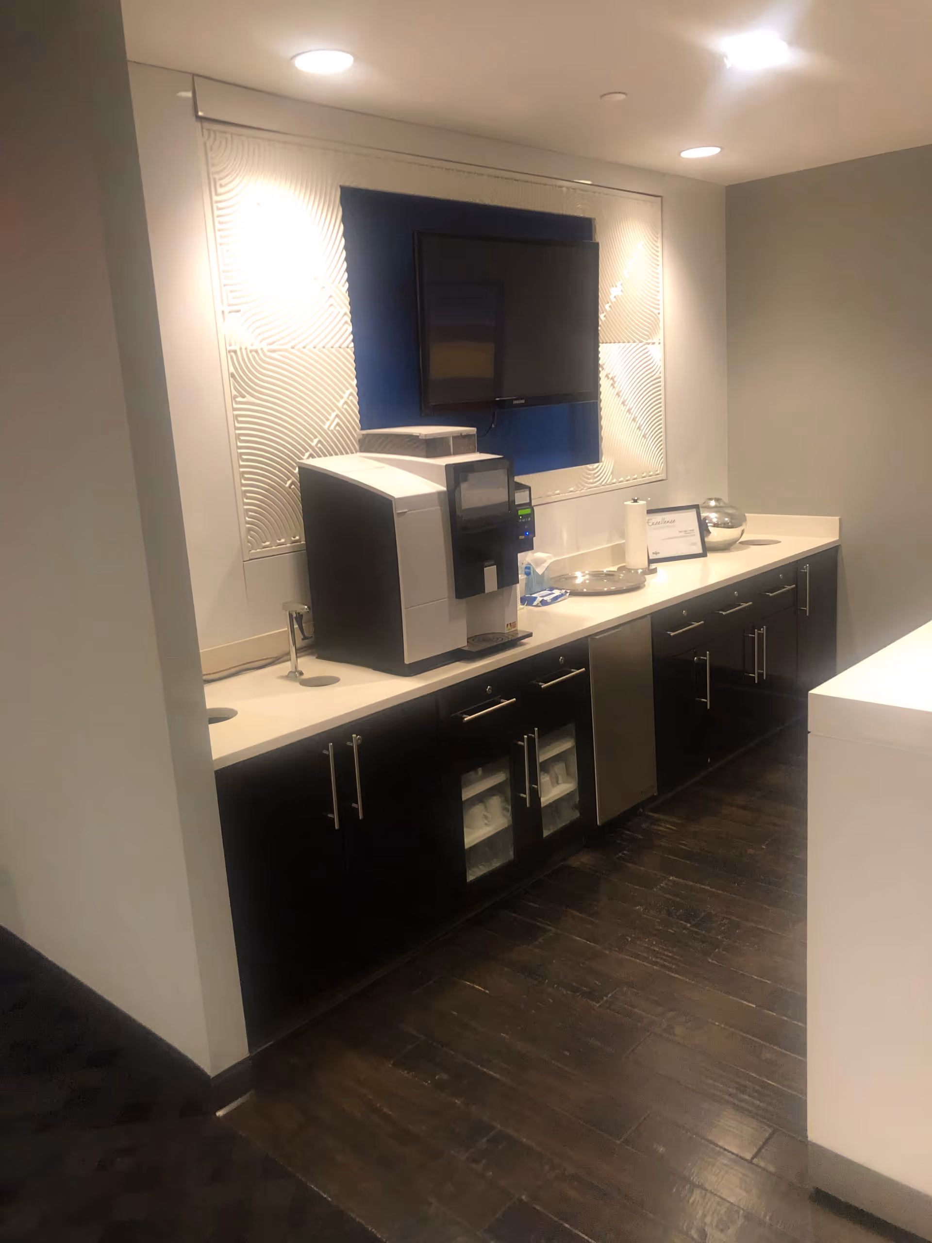 A modern kitchen area with a coffee machine, cabinets, a small fridge, and a wall-mounted TV.