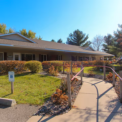 Front exterior of a single-story assisted living building with a ramped concrete walkway and handrails, landscaped lawn and shrubs under a clear blue sky.