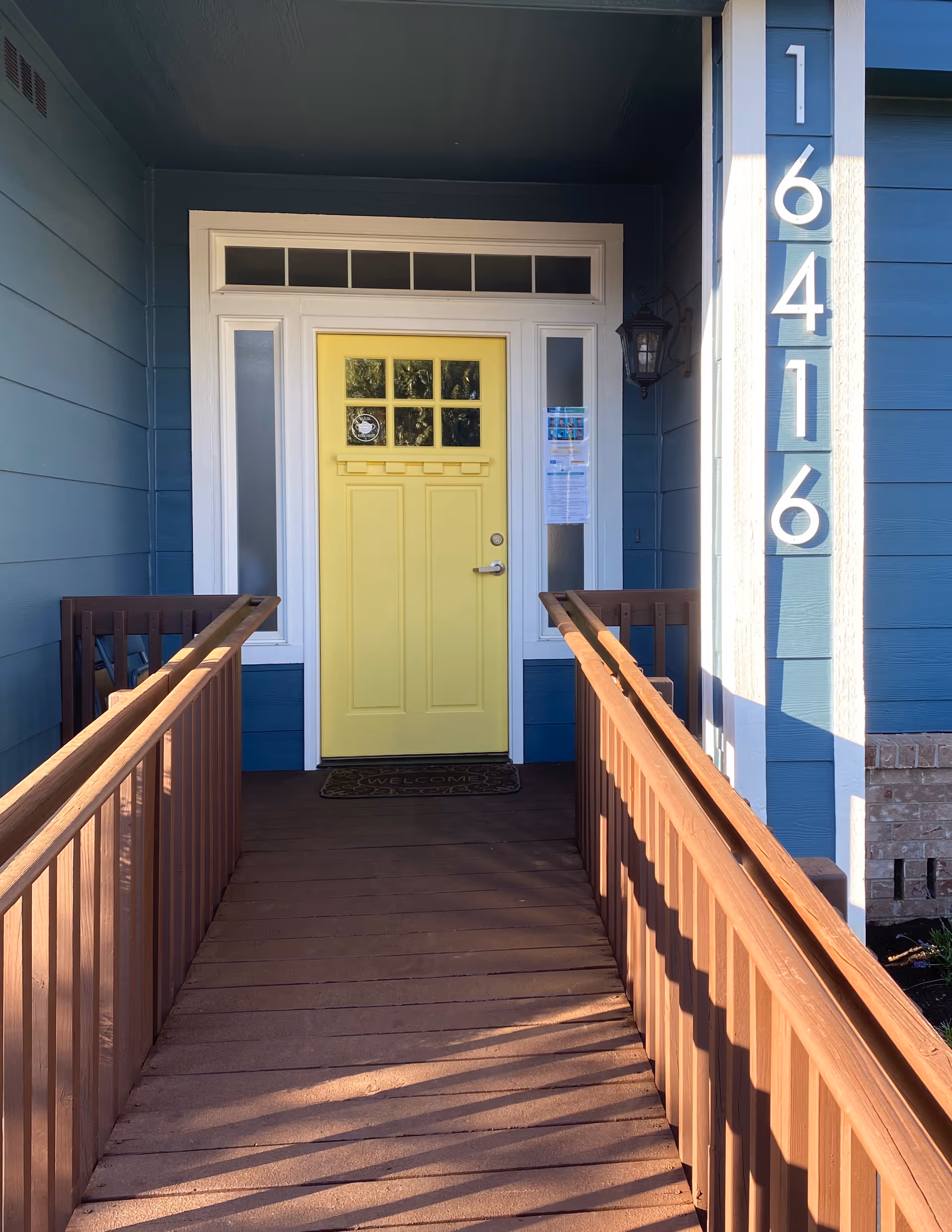 Blue building entry with a yellow front door, wooden ramp, and numbered vertical columns.