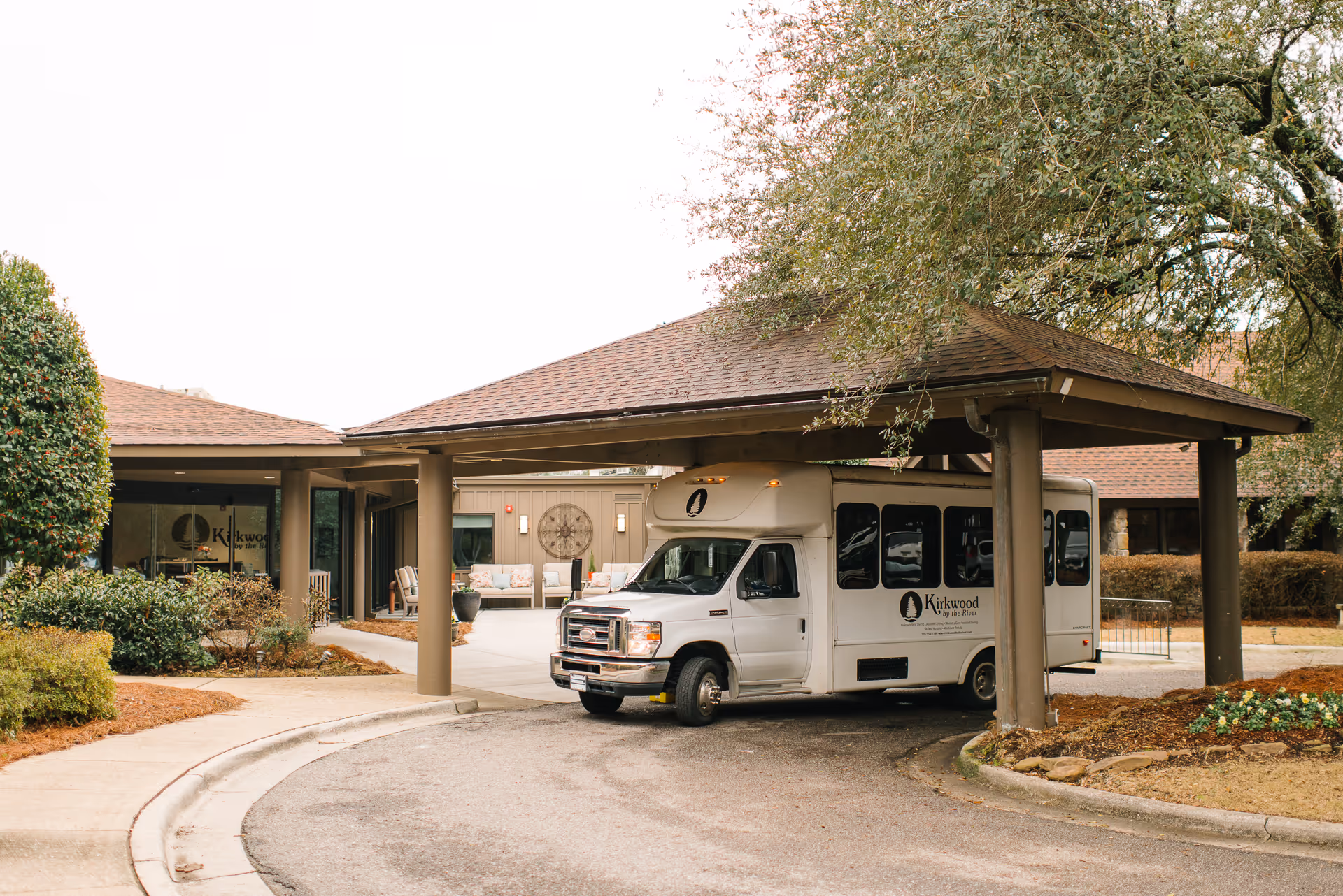 A white shuttle bus parked under a covered driveway at the entrance of a senior living facility named Kirkwood By the River. The building has a brown roof and beige walls, with some outdoor seating visible near the entrance. There are trees and landscaped bushes around the driveway.