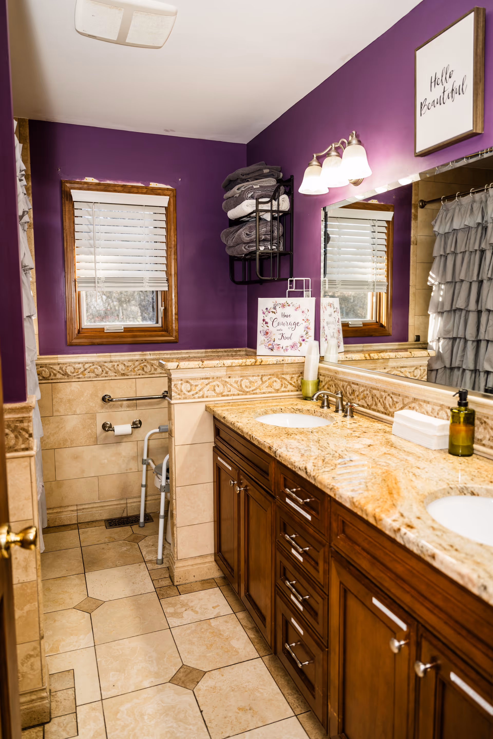 Bathroom with purple walls, a double-sink granite countertop and wooden vanity, wall towel rack, window, and a shower with ruffled curtain.