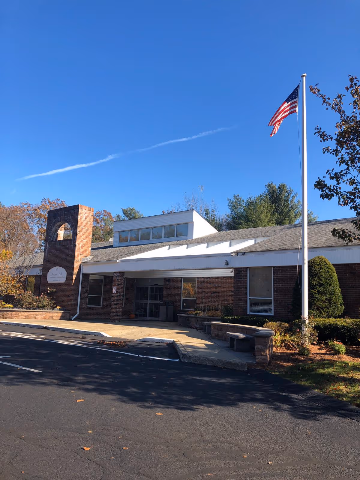 Front entrance of a single-story brick rehabilitation and healthcare building with an American flag on a tall pole against a clear blue sky.
