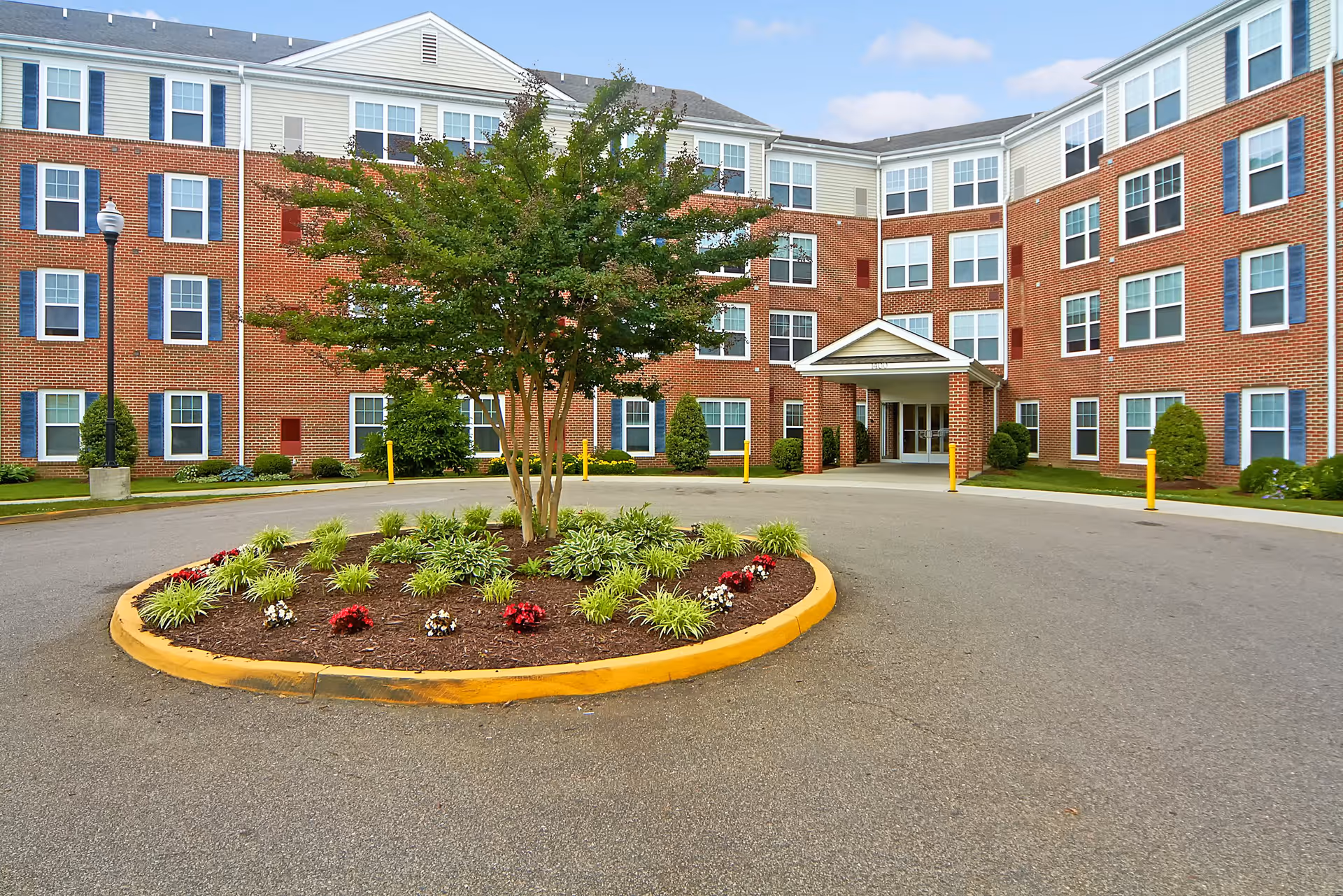 Exterior view of a multi-story brick senior living facility with numerous windows, a circular driveway with a landscaped island featuring a tree and flowers, and a covered entrance.