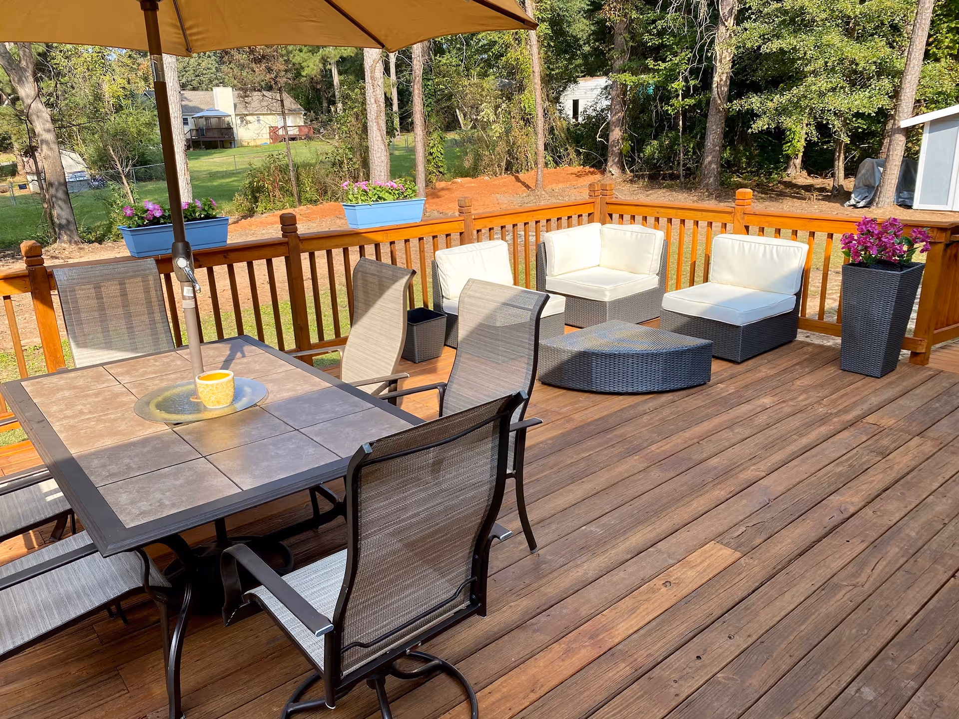 Sunlit wooden deck with patio furniture, an umbrella, and planters in a spacious yard.