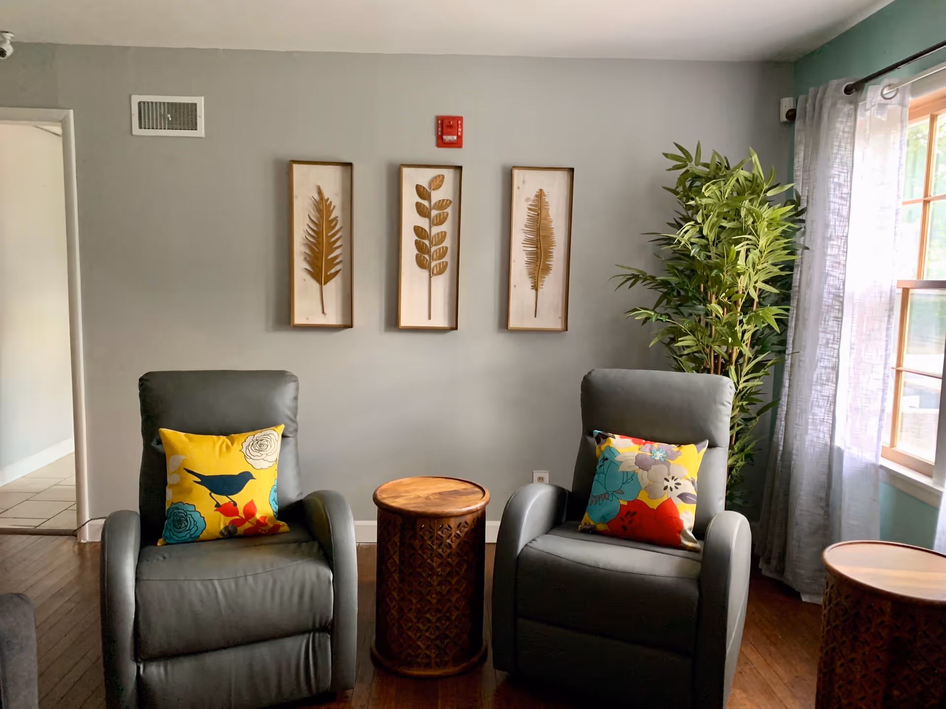 A cozy living room area with two gray recliner chairs, each adorned with colorful floral and bird-patterned throw pillows. Between the chairs is a small round wooden side table with a carved design. On the wall behind the chairs are three framed botanical prints featuring different leaf designs. To the right, a large window with sheer white curtains lets in natural light, and a tall green potted plant stands next to the window. The room has wooden flooring and light gray walls.