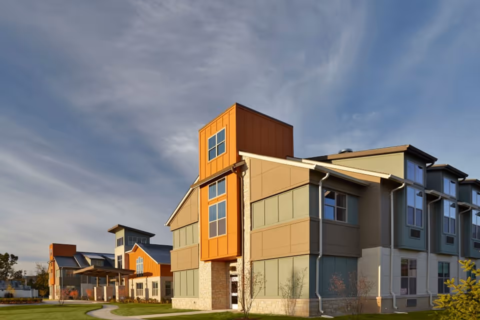 Front exterior of a modern multi-story senior living building with orange and beige siding under a blue sky.