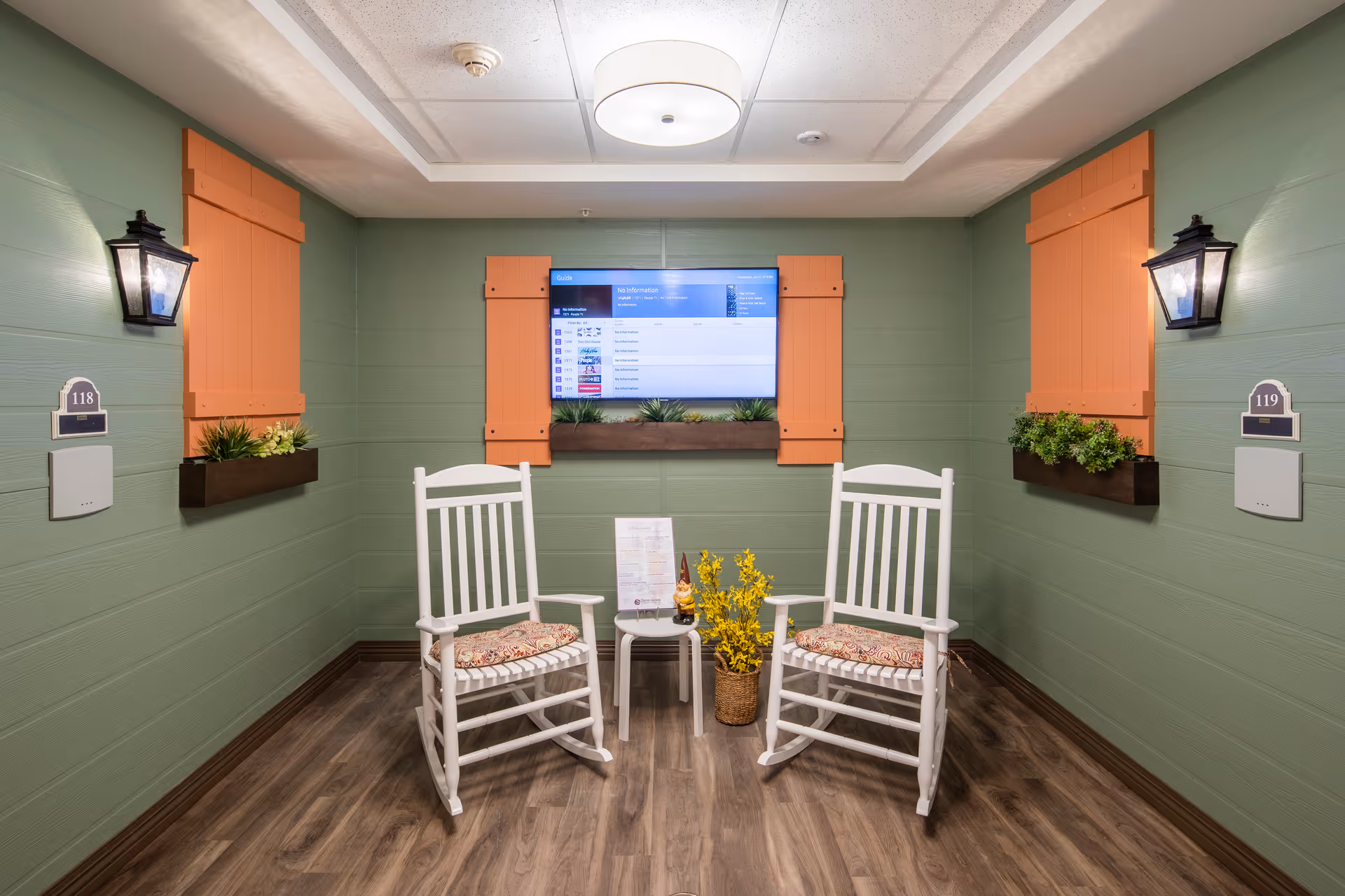 Small interior seating area with two white rocking chairs facing a wall-mounted TV, orange decorative shutters, wall lanterns and potted plants.