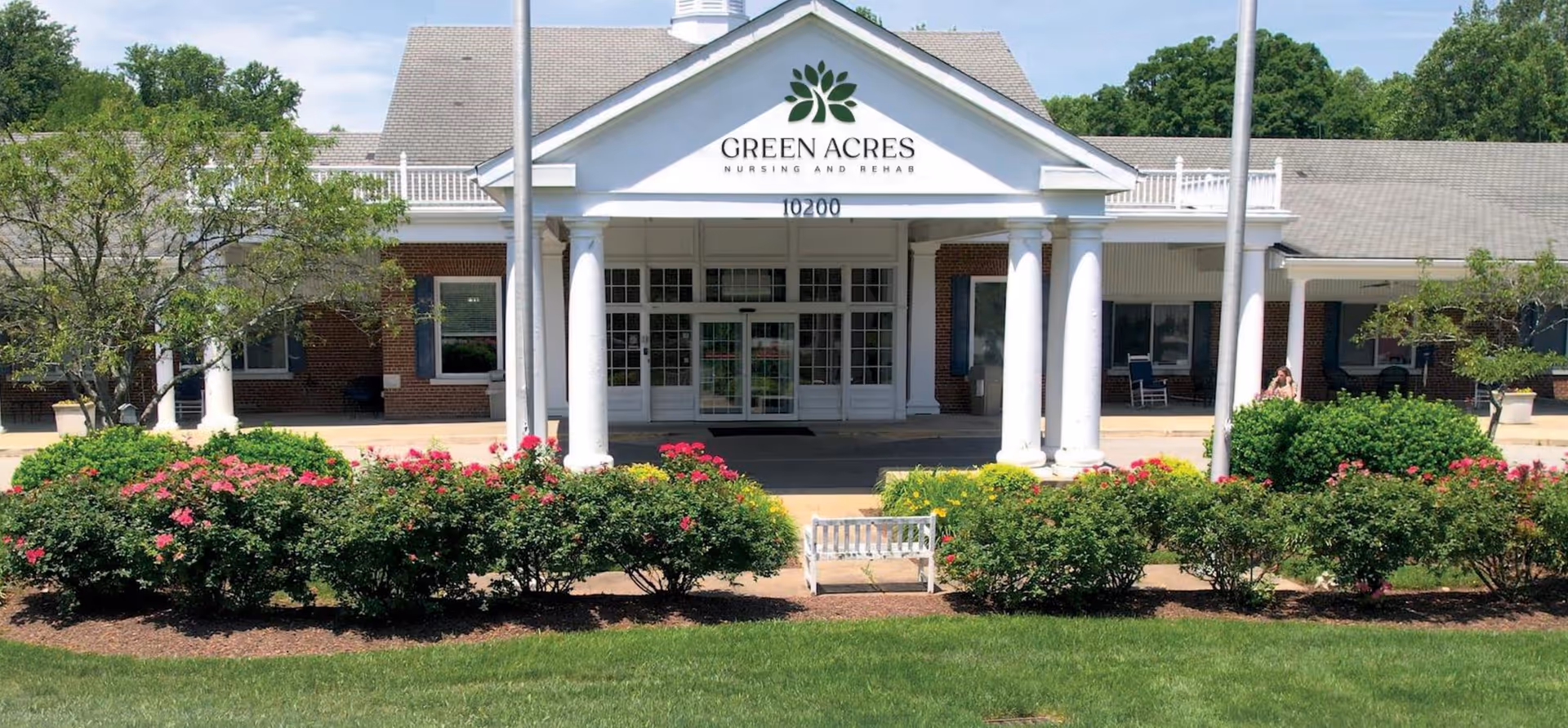 Front entrance of a nursing facility with white columns, greenery, and a bench.