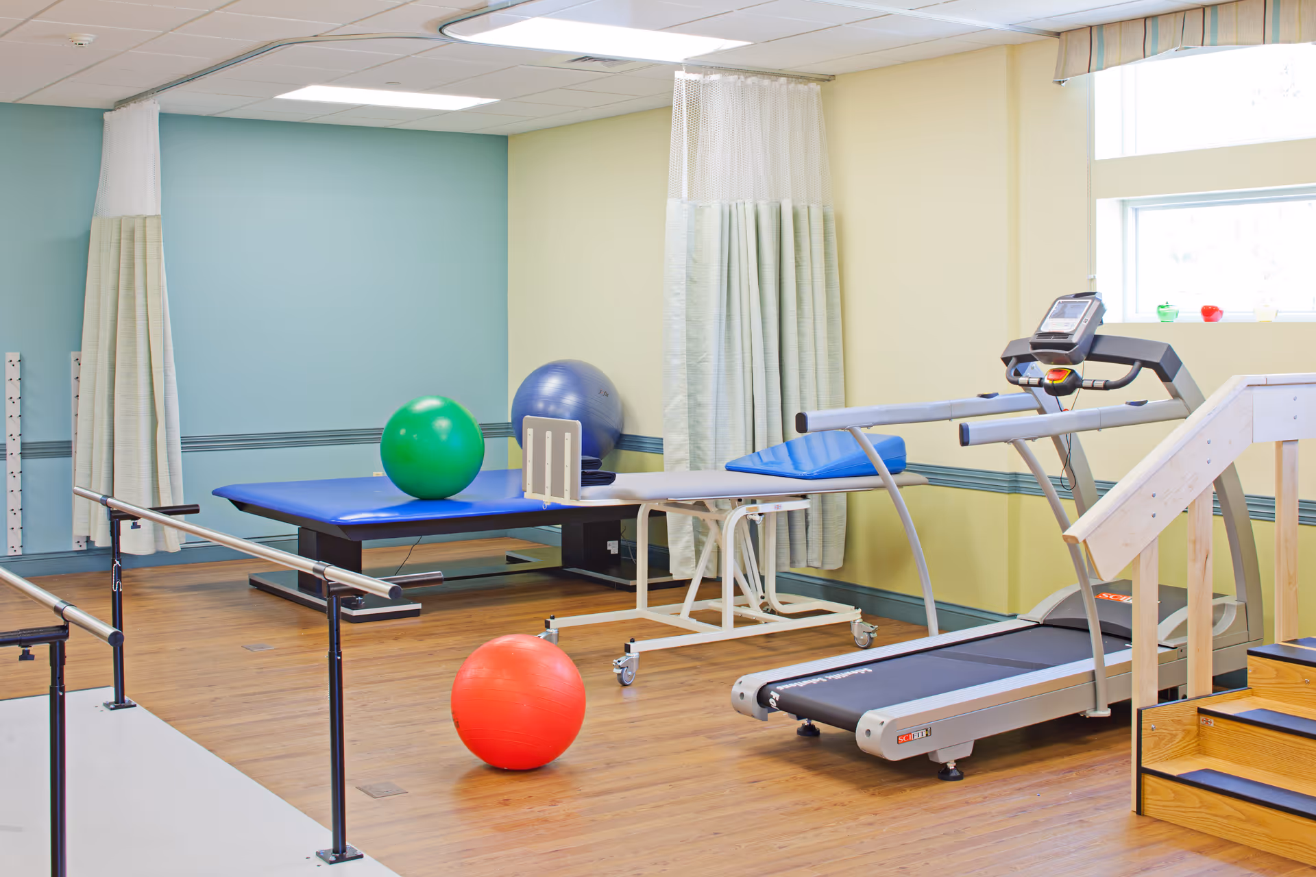 A physical therapy or rehabilitation room with a treadmill, parallel bars for walking exercises, therapy tables, and colorful exercise balls. The room has light blue and yellow walls, wood flooring, and a window with a valance.