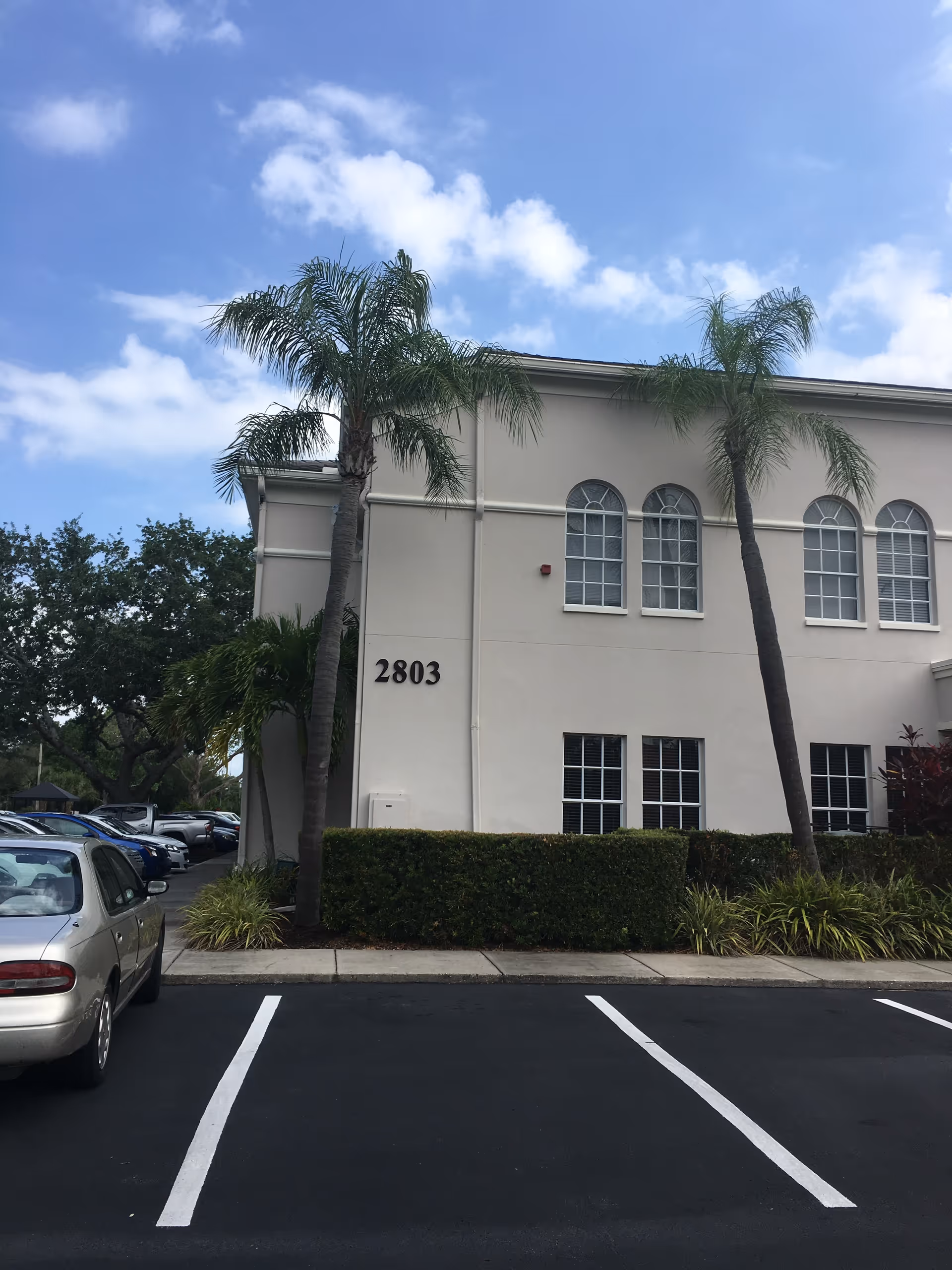 Beige senior living building with arched windows, palm trees, and a parking lot in front