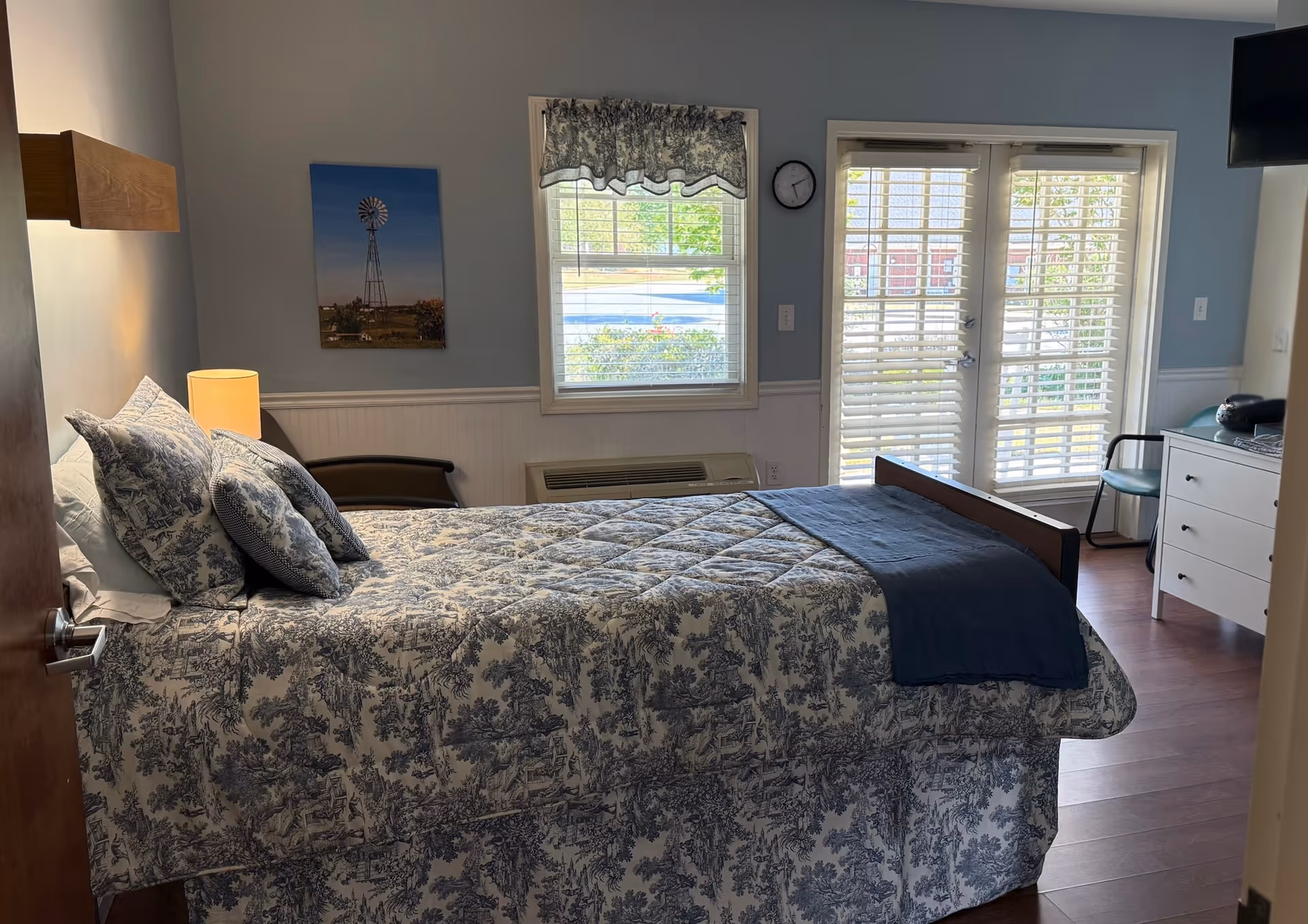 A tidy bedroom with a blue patterned bedspread, windows, and French doors.