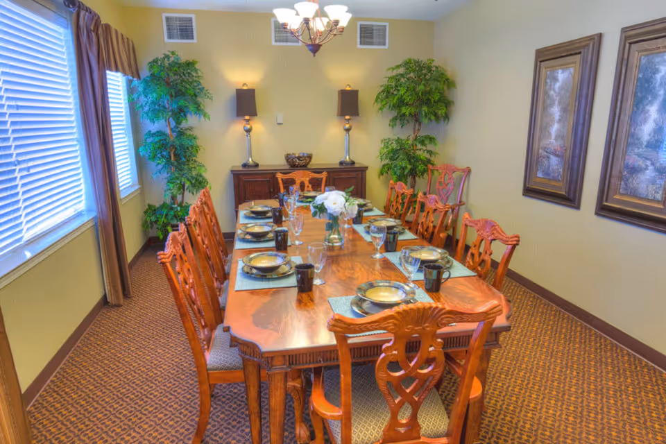 A formal dining room with a long wooden table set for eight people, featuring plates, bowls, glasses, and black mugs. The room has patterned carpet, two large green plants in the corners, two framed paintings on the wall, a wooden sideboard with two lamps, and a chandelier overhead. Large windows with blinds and curtains are on one side of the room.