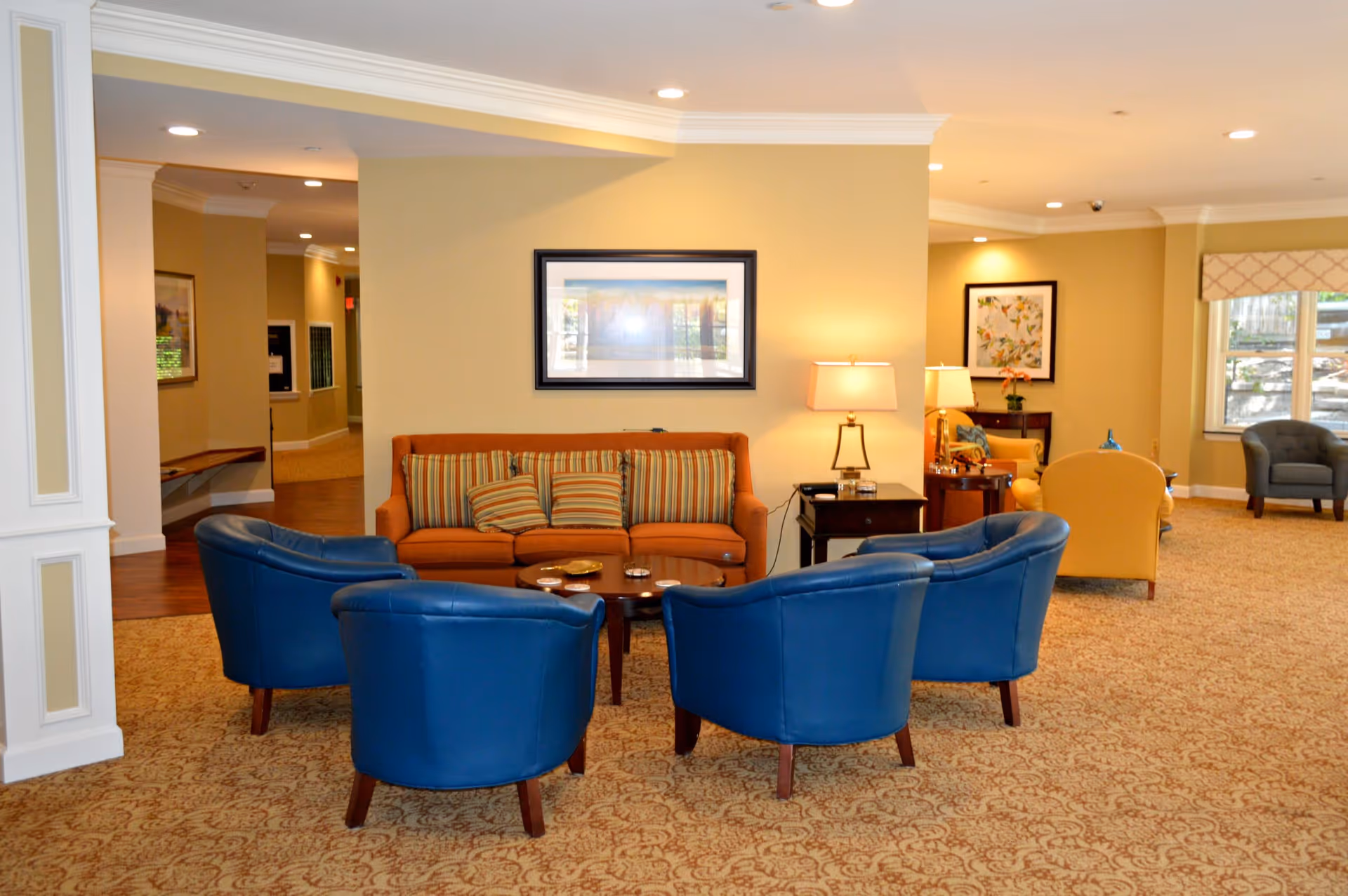 A cozy living room area in a senior living facility with a brown sofa adorned with striped cushions, surrounded by four blue armchairs arranged around a wooden coffee table. The room has beige walls with framed artwork, a side table with a lamp, and carpeted flooring. Additional seating and windows are visible in the background.