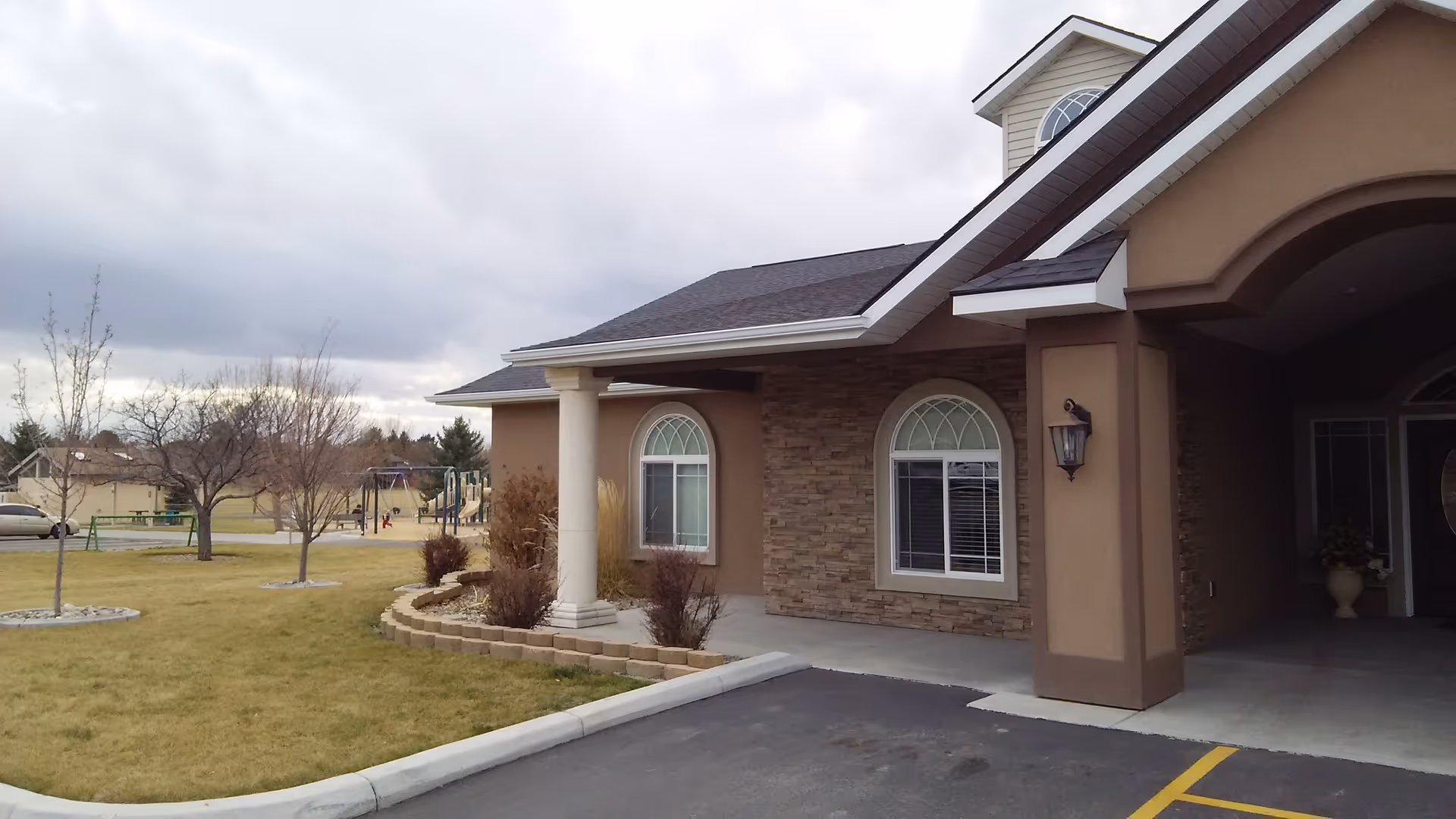 Front entrance of a beige building with arched windows, white columns, a covered driveway and lawn with a distant playground.