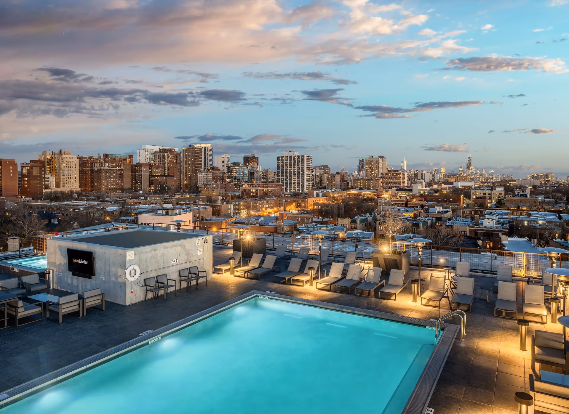 Rooftop swimming pool with lounge chairs and city skyline at dusk.